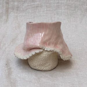 Puede incluir: Un jarrón de cerámica hecho a mano con un diseño único en forma de hongo. La parte superior es de color rosa claro con un borde ondulado, y la base es de color blanco roto texturizado. El jarrón tiene un acabado brillante y está sobre un fondo neutro.