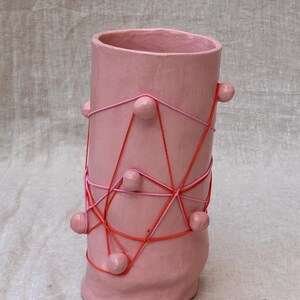 Puede incluir: Un jarrón de cerámica rosa con forma cilíndrica y base redondeada. El jarrón está adornado con cuerdas rosas y naranjas, y pequeñas esferas rosas están adheridas. El jarrón tiene un diseño artístico único.
