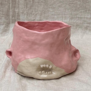 Puede incluir: Una maceta de cerámica rosa con un diseño texturizado único. La maceta presenta un exterior rosa claro y una base blanca crema contrastante con pequeños dientes blancos. Dos pequeñas asas están en los lados. Esta pieza hecha a mano añade un toque de fantasía a cualquier espacio.