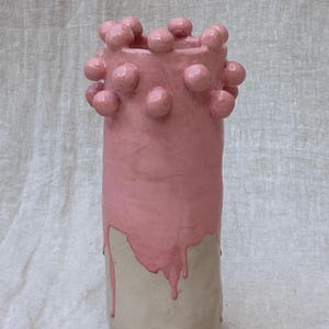 Puede incluir: Un jarrón de cerámica hecho a mano con forma cilíndrica. La parte superior está pintada de color rosa claro con pequeños detalles redondos en relieve. La pintura rosa gotea sobre la base sin pintar, de color blanquecino. Un objeto decorativo.