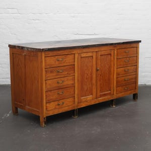 Puede incluir: Un mueble de madera antiguo con una parte superior oscura y numerosos cajones y puertas. El mueble tiene un tono de madera cálido y natural con vetas visibles y herrajes de latón. Cuenta con múltiples cajones a cada lado y un armario central con puertas.