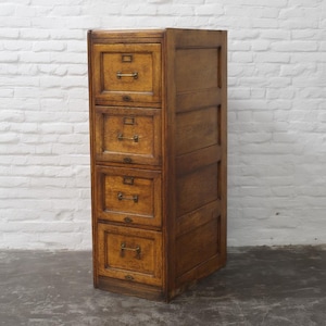 May include: A vintage wooden file cabinet with four drawers. The cabinet is a warm brown color with brass hardware. The drawers have rectangular pulls and small keyholes. The cabinet is against a white brick wall.