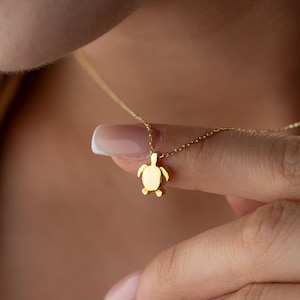 May include: A gold-toned necklace featuring a small, smooth turtle-shaped pendant suspended from a delicate, thin chain. The pendant has a polished, reflective surface and is shaped like a sea turtle with visible flippers and a rounded shell.