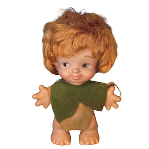 May include: A vintage doll with short, reddish-brown hair, wearing a green felt vest and a brown cloth diaper. The doll has a painted face with brown eyes and a smile.