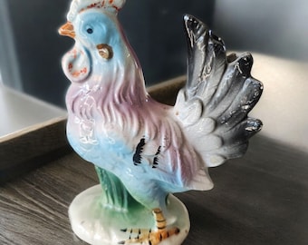 May include: A colorful ceramic rooster figurine with a blue, pink, and white body, a black tail, and a red comb. The rooster is standing on a green base with a white rim.