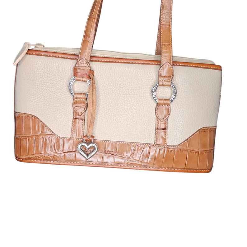 May include: Cream-colored handbag with brown crocodile-textured accents. Features silver-toned hardware, a heart-shaped charm, and dual top handles. The bag has a zippered top closure and a structured design.