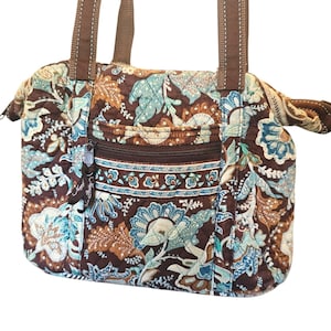 May include: A quilted handbag featuring a brown, light blue, and cream floral paisley pattern. The bag includes two brown fabric shoulder straps, a zippered front pocket with a dark brown zipper pull, and a main zippered compartment closure.