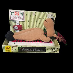 May include: A plush reindeer toy in a box, designed for Christmas. The reindeer is lying down on a red and white striped surface. The box has the text "Exercise Reindeer" and "25 More Days Til Christmas".