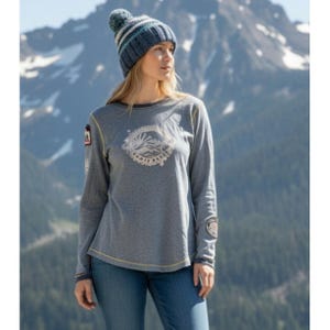 May include: A woman wearing a blue knit beanie with white and blue stripes, a gray long-sleeve shirt with a mountain graphic, and blue jeans. The shirt has patches on the sleeves and a yellow trim.