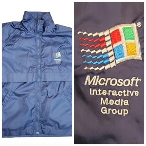 May include: A navy blue windbreaker jacket with a zipper closure and two pockets. The jacket has an embroidered Microsoft Interactive Media Group logo on the front. The logo features the Microsoft Windows logo and the text "Microsoft Interactive Media Group".