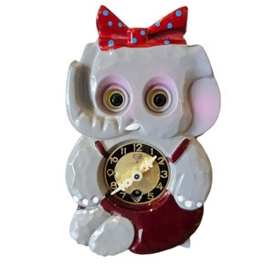 May include: A gray ceramic elephant clock with a red bow and a clock face with black numbers. The clock has a red and white striped dress and a white polka dot bow.