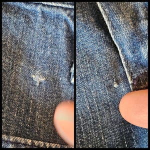 May include: Close-up of two sections of blue denim fabric. The fabric has a textured appearance with small, light-coloured flecks. A finger is visible in the lower right corner of the second section, providing scale. The denim appears to be from a jacket or trousers.