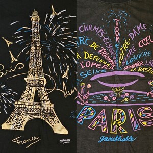 May include: A black t-shirt with a gold Eiffel Tower graphic and fireworks, the words "Paris" and "France" are written in gold script. The right side of the shirt has a colorful graphic of a fountain with the words "Paris" and "Inoubliable" written in colorful script.