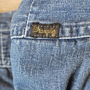 May include: Close-up of blue denim fabric. A black rectangular label with the gold-coloured word "Wrangler" is stitched onto the denim. The label is bordered by a thin, dark thread.