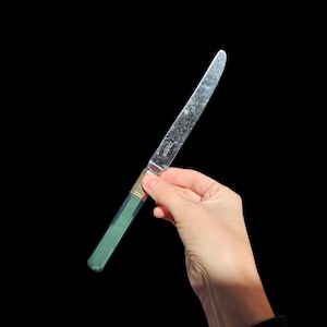 May include: A silver butter knife with a green handle and gold accents. The knife is held against a black background. The blade is slightly tarnished, showing signs of use. The handle is a solid, opaque green color.