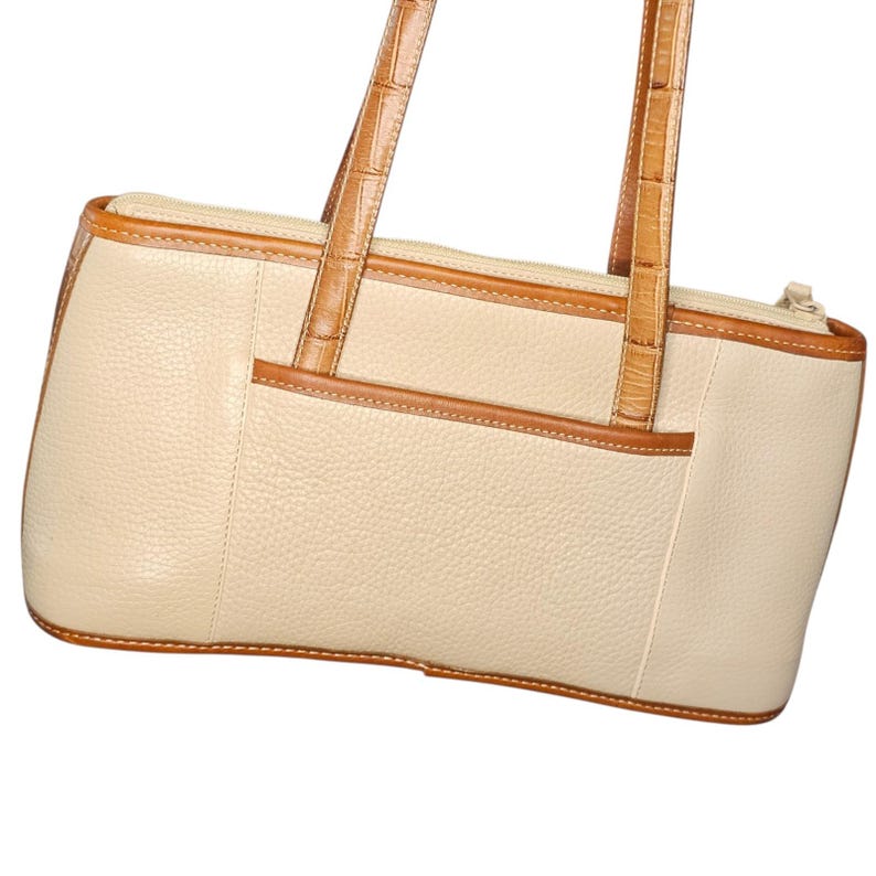 May include: Cream-colored leather tote bag with brown trim and handles. The bag has a rectangular shape with a zipper closure and a small pocket on the front. The handles are a lighter brown with a textured pattern.