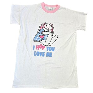 May include: White t-shirt with a pink ringer neck. The shirt features a cartoon bunny wearing a blue and white striped dress and holding a pink heart. The text "I HOP YOU LOVE ME" is printed below the bunny.