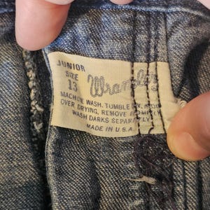 May include: Close-up of a denim garment label. The label reads "JUNIOR SIZE 13 Wrangler" with washing instructions. The label is white with black text and is sewn into the blue denim fabric. "MADE IN USA" is printed on the label.