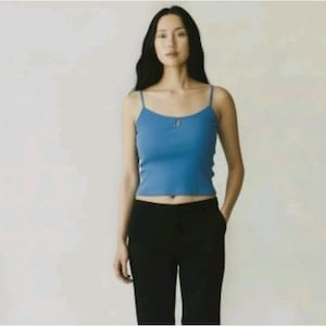 May include: A woman wearing a blue ribbed tank top with thin straps and a small keyhole detail. She is also wearing black pants. The background is a plain, light-colored wall.