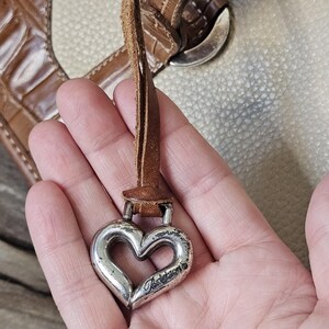 May include: Silver heart-shaped charm with the word "Brighton" engraved on it, attached to a brown leather strap. The charm is held in front of a handbag with a brown and cream color scheme.