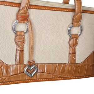 May include: Cream-colored handbag with brown crocodile-textured accents. Features silver-tone hardware, including decorative rings and a heart-shaped charm. The bag has a top zipper and brown leather handles.