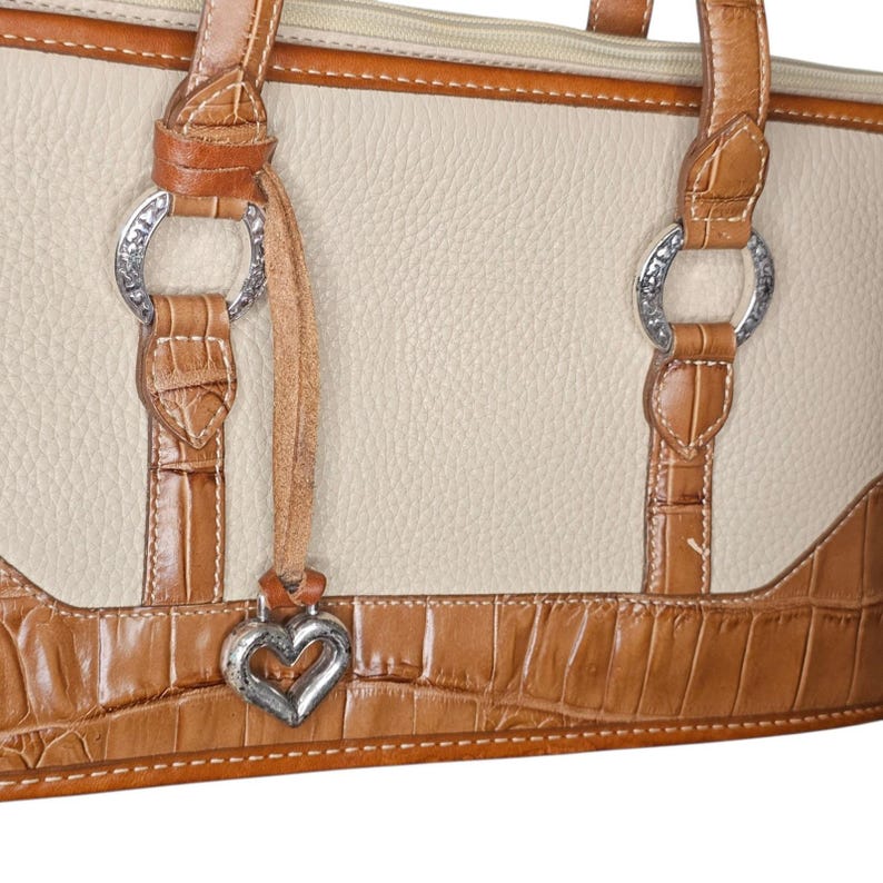 May include: Cream-colored handbag with brown crocodile-textured accents. Features silver-tone hardware, including decorative rings and a heart-shaped charm. The bag has a top zipper and brown leather handles.
