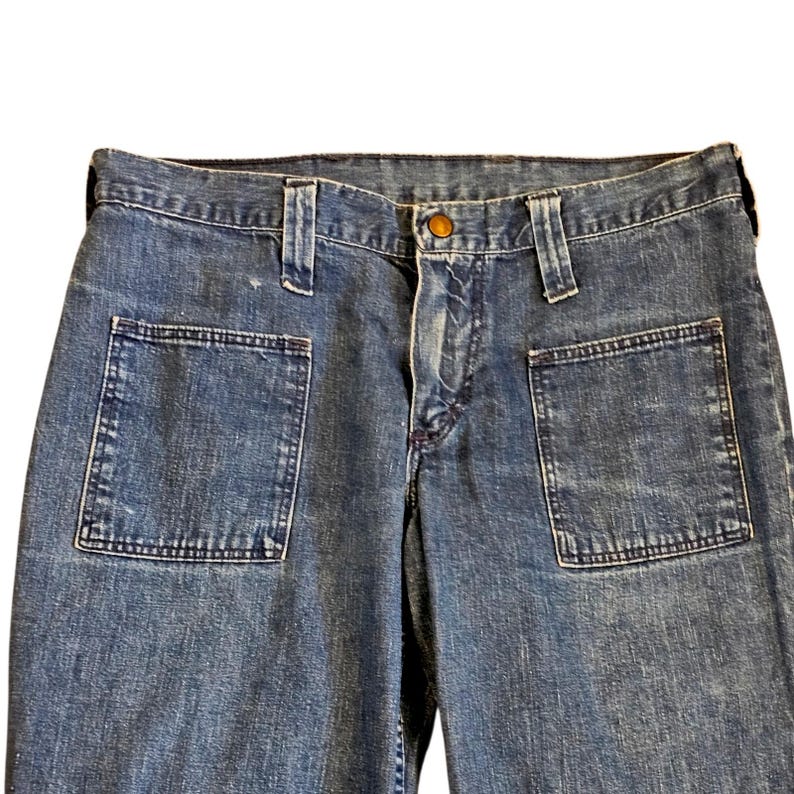 May include: Close-up of blue denim jeans with large square front pockets. The jeans have a button closure and belt loops. The denim has a faded appearance.