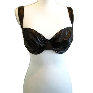 May include: A black bra with a shiny, textured appearance. The bra has underwire support and adjustable straps. The straps and cups are connected by a thin band. The bra is displayed on a white mannequin.