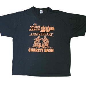 May include: Black t-shirt with orange text and graphics. The text reads "The Central Illinois ABATE 20th Anniversary Charity Bash." The graphic depicts two motorcyclists. The shirt is a vintage item.
