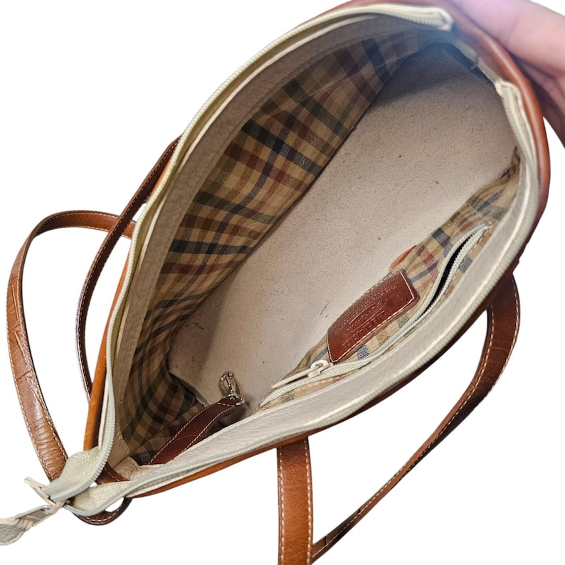 May include: Open view of a brown leather tote bag with a cream-colored interior. The bag features a plaid lining and a zippered pocket. The handles are also brown leather.