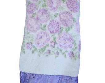 May include: A white and purple floral throw blanket with a purple satin trim. The blanket features a repeating pattern of purple roses on a white background.