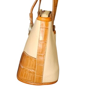 May include: Cream-colored handbag with brown crocodile-textured accents. The bag features a structured design with a rounded base and a top handle. The trim and handle are a lighter shade of brown.