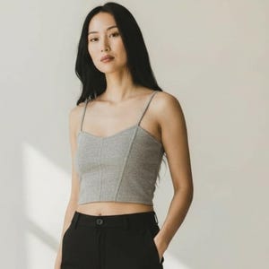 May include: A gray crop top with thin straps and a sweetheart neckline. The top is paired with black pants. The model is standing in front of a white background.