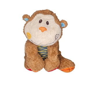 May include: A plush monkey toy with brown fur, a cream-colored face, and blue accents on the ears. The monkey wears a blue and yellow striped shirt. The toy has orange and red details on its feet.