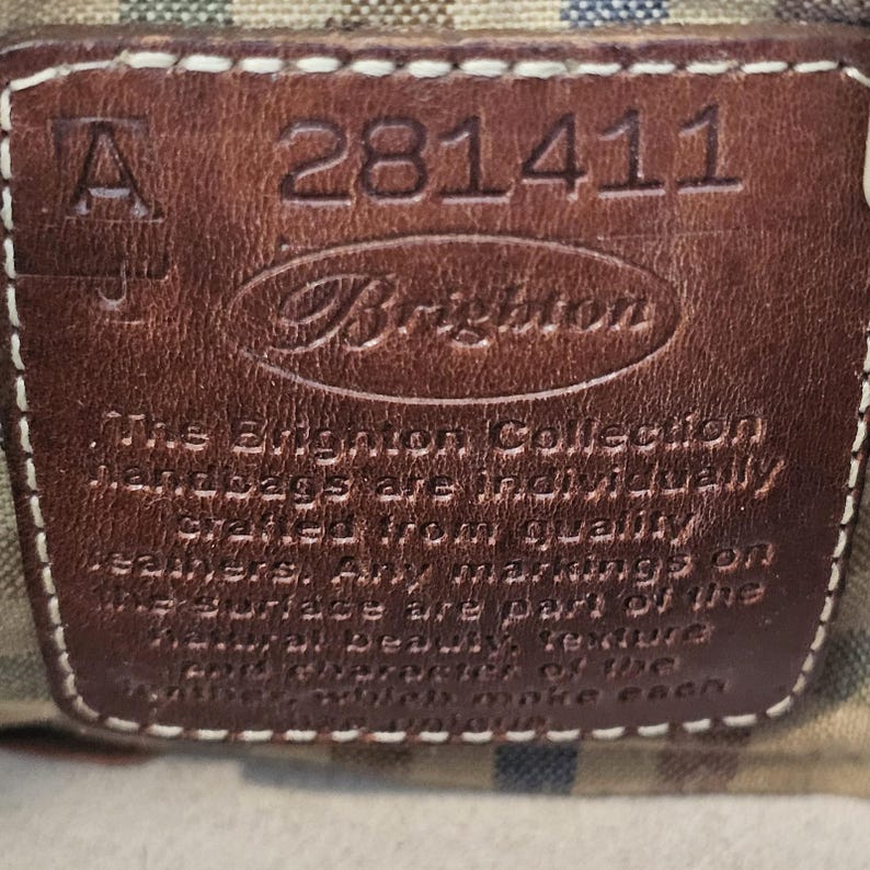 May include: Close-up of a brown leather tag with the Brighton logo and the number 281411. The tag is stitched with white thread and includes text that reads, "The Brighton Collection handbags are individually crafted from quality leathers."