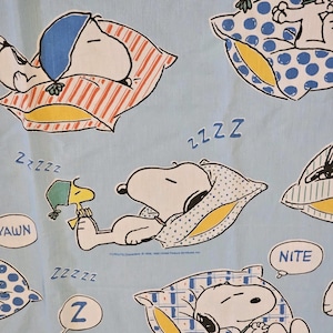 May include: A light blue fabric featuring a repeating pattern of Snoopy and Woodstock sleeping on various pillows. The design includes blue, yellow, and white accents, with text bubbles containing the words YAWN and NITE, and ZZZZZ symbols.