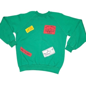 May include: Green sweatshirt with patches featuring text and illustrations. Patches include "Galway Bay", "Trinity College Dublin", "Lakes of Killarney", and "Hills of Donegal". The sweatshirt has a crew neck and long sleeves.