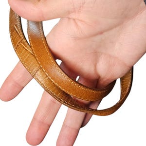 May include: Close-up of a tan leather handbag strap. The strap is a light brown color with a textured surface and white stitching along the edges. The strap is held by a hand against a white background.