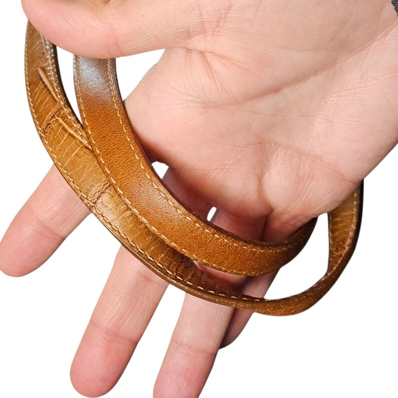 May include: Close-up of a tan leather handbag strap. The strap is a light brown color with a textured surface and white stitching along the edges. The strap is held by a hand against a white background.