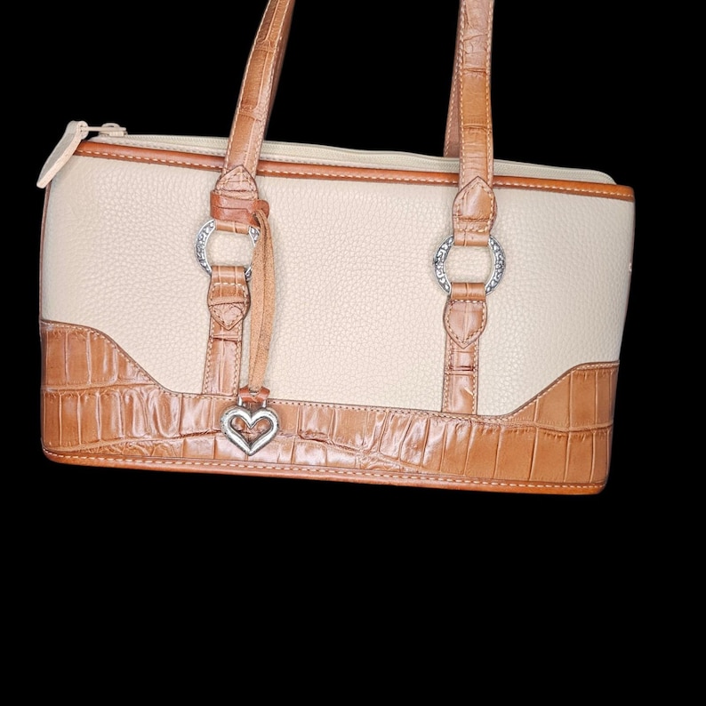 May include: Cream-colored handbag with brown leather accents and handles. Features a textured, faux-crocodile pattern on the bottom and a silver heart-shaped charm. The bag has a zipper closure and silver hardware.