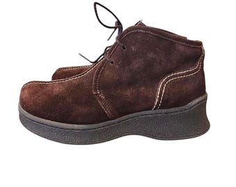 May include: Brown suede ankle boots with a lace-up design and a thick black sole. The boots have white stitching details and a rounded toe. These casual boots are suitable for everyday wear.