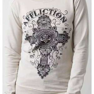 May include: A cream-colored long-sleeved sweatshirt with a black and white graphic print of a cross with the word "AFFLICTION" written around it.