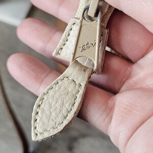 May include: Close-up of a beige zipper pull with the brand name "Talon" embossed on the metal zipper head. The pull tab is made of stitched leather. The zipper is a neutral color.