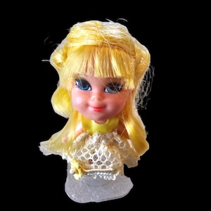 May include: A vintage doll head with yellow hair and blue eyes. The doll has a yellow collar and a white lace top. The doll is mounted on a clear plastic base. The background is black.