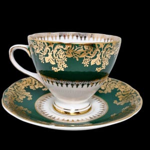 May include: A green and gold teacup and saucer set. The teacup and saucer are decorated with gold floral patterns and trim. The teacup has a white interior and a curved handle. The saucer is round and has a matching design.
