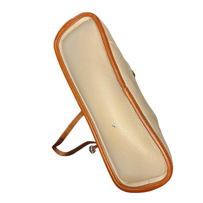 May include: Cream-colored handbag with brown leather trim and a matching strap. The bag has a textured surface and a rectangular shape. The strap is attached to the bag with silver-colored hardware.