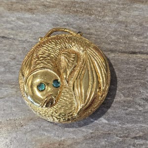 May include: A gold-toned, circular brooch featuring a stylized fish design. The fish has teal-colored eyes and detailed scales. The brooch has a loop at the top and is set against a textured background.
