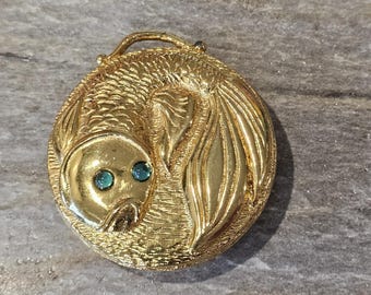 May include: A gold-toned, circular brooch featuring a stylized fish design. The fish has teal-colored eyes and detailed scales. The brooch has a loop at the top and is set against a textured background.