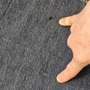 May include: Close-up of dark blue denim fabric with a small dark stain. The fabric has a textured appearance with visible diagonal lines. A hand is pointing towards the stain.