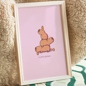 May include: A framed print with a cartoon illustration of three capybaras balancing on top of each other, with a small orange fruit on top. The print has a pink background and the text "STACKYBARA" at the bottom.